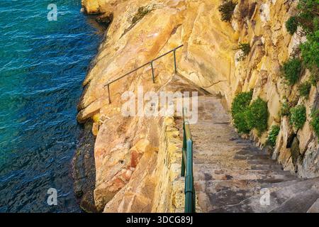 Escaliers étroits en pierre avec des garde-corps en métal sur une falaise de calcaire menant aux eaux bleues claires de la Méditerranée. Banque D'Images