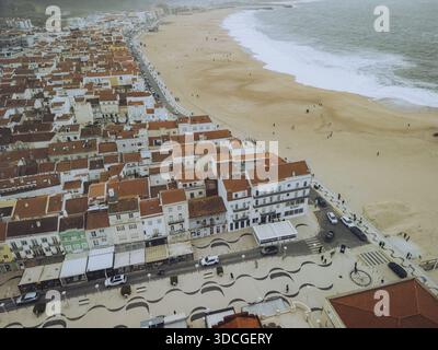 Vue aérienne des toits en terre cuite descendant en cascade pour rencontrer les sables dorés où les puissantes vagues de l'Atlantique s'écrasent, Nazaré, district de Leiria, Portugal. Banque D'Images