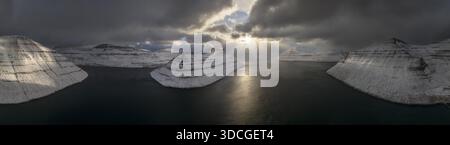 Vue aérienne de rayons de soleil perçant à travers des nuages sombres au-dessus de montagnes couvertes de neige, en terrasses et de l'eau calme, sombre, Vagar, Vagar, îles Féroé. Banque D'Images
