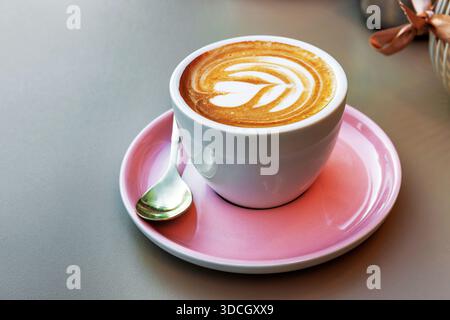 Une tasse de cappuccino avec une fleur de coeur en forme de latte art sur une soucoupe rose avec une petite cuillère sur une surface de table lisse. Banque D'Images