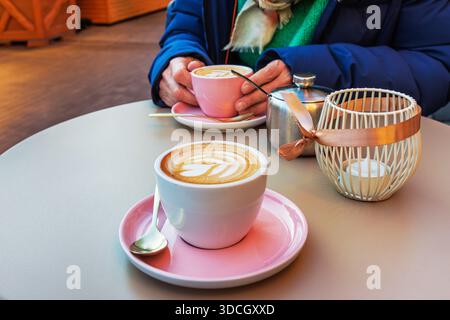 Deux tasses de café avec de l'art latte assis sur une table de café avec une lanterne de bougie et une personne tenant la tasse en arrière-plan. Banque D'Images