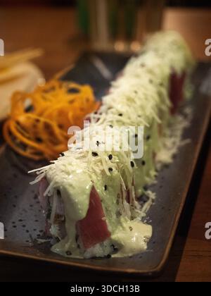 Une photographie en gros plan montre un long rouleau de sushi sur une assiette rectangulaire sombre. Le rouleau est généreusement garni d'une sauce crémeuse vert clair, râpée Banque D'Images