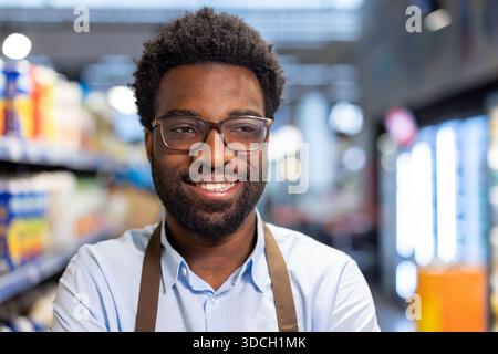 Jeune homme afro-américain avec des lunettes et un tablier souriant en toute confiance tout en travaillant dans un supermarché, représentant un excellent service à la clientèle et un personnel de vente au détail amical Banque D'Images