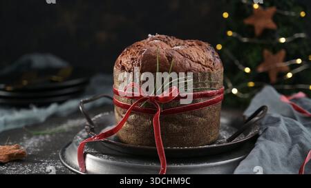 Un grand panettone classique est enveloppé dans un noeud en velours rouge et présenté sur un plateau en métal vintage. Le décor festif et démodé présente des lumières de fée dorées, Banque D'Images
