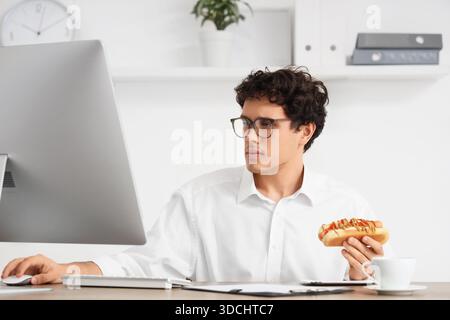 Jeune homme d'affaires avec hot dog savoureux travaillant dans le bureau Banque D'Images