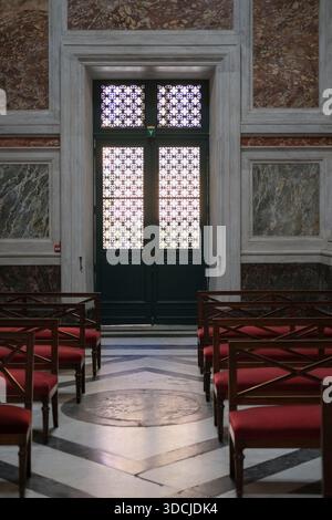 Vue intérieure symétrique montrant des rangées de sièges en bois, des chaises rembourrées rouges, des panneaux muraux en marbre, un sol en pierre à motifs, des portes vertes fermées Banque D'Images
