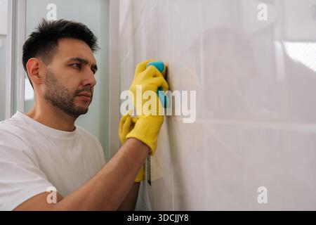 Homme barbu concentré nettoyant les carreaux de salle de bain avec une éponge bleue tout en portant des gants en caoutchouc jaune, en se concentrant sur le nettoyage du mur carrelé blanc pour Banque D'Images