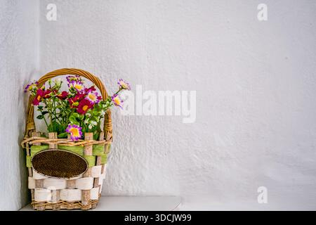 Un panier tissé rempli de fleurs colorées se trouve dans un coin contre un mur blanc. Banque D'Images