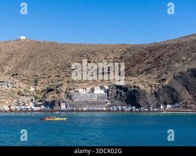 Vue sur une plage animée avec des parasols colorés et des gens profitant du soleil sur fond de falaises accidentées et un ciel bleu clair, Oia, Thira, Gree Banque D'Images