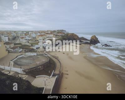 Vue aérienne d'une plage tranquille rencontrant la côte accidentée, dominée par le phare blanchi à la chaux et la ville de Santa Cruz, Santa Cruz, Lisboa, Banque D'Images