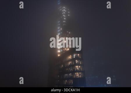 Gratte-ciel futuriste enveloppé dans un brouillard nocturne dense. prise de vue cinématographique en bas angle d'un gratte-ciel en verre moderne traversant une épaisse brume nocturne. Incandescence de Banque D'Images