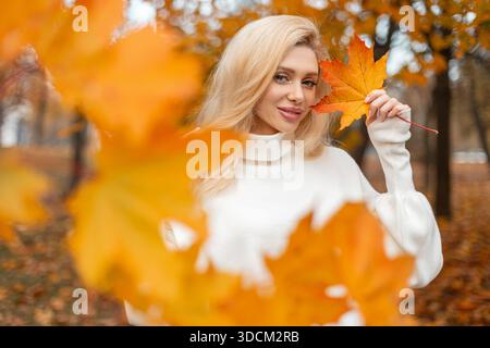 Femme attrayante dans une tenue confortable profitant de la nature pendant la saison d'automne. Positive Trendy Girl tient une feuille d'érable jaune coloré brillant. Humeur automnale et Banque D'Images