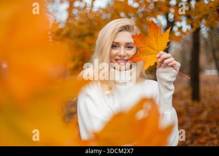 Femme blonde souriante dans un pull blanc tenant une feuille d'érable d'automne devant son visage, debout dans un parc avec foli d'automne orange et jaune vif Banque D'Images