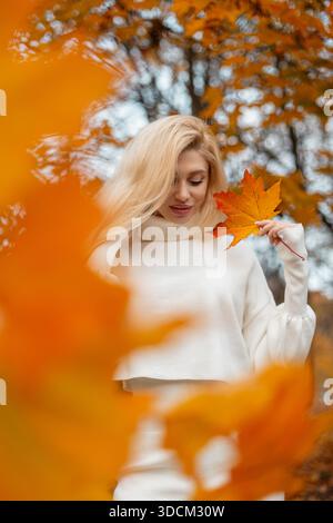 Femme blonde souriante en pull blanc tenant une feuille d'érable dans le parc d'automne avec feuillage doré. Positive Trendy Girl détient une carte jaune colorée brillante Banque D'Images