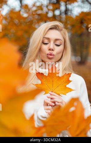 Femme embrasse et tient une grande feuille d'érable dans la forêt d'automne dorée. Positive Trendy Girl tient une feuille d'érable jaune coloré brillant. Humeur automnale et vif Banque D'Images
