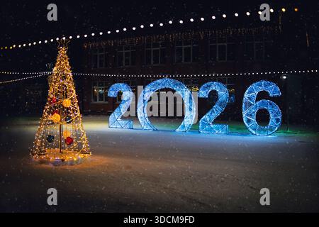 Description : une enseigne numérique éclairée ou une installation affichant 2026 dans une rue enneigée la nuit, décorée de guirlandes festives lors d'un fal de neige hivernal Banque D'Images