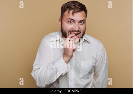 Un homme se tient debout dans un studio. Il sourit et touche sa bouche avec son doigt. Il porte une chemise blanche. L'arrière-plan est de couleur unie. La lumière est chaude Banque D'Images