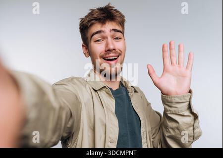 Un jeune homme se tient devant un fond Uni. Il sourit et agite devant la caméra pendant qu'il prend un selfie. Son expression montre le bonheur et l'ene Banque D'Images