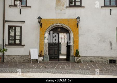 Un tunnel en pierre voûtée encadre une vue sur un bâtiment historique blanc avec des volets bruns et une fresque religieuse fanée. Une échelle en bois s'accroche Banque D'Images