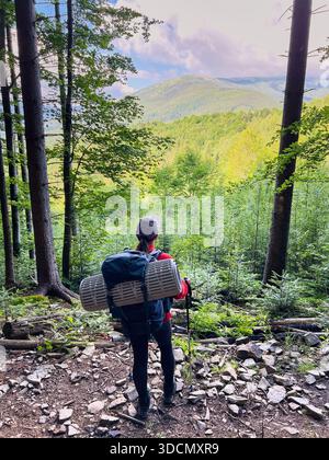 Vue arrière du randonneur avec grand sac à dos et matelas sur le sentier de la forêt rocheuse, contemplant la vue panoramique sur la montagne. Une végétation luxuriante et de grands arbres encadrent des collines lointaines et vallonnées sous un ciel partiellement nuageux. Banque D'Images