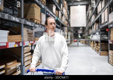 Jeune client masculin poussant un chariot dans une allée d'entrepôt de meubles, regardant des boîtes empilées sur des étagères hautes. Photo de haute qualité Banque D'Images