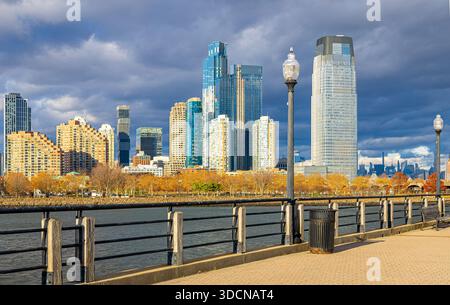 Horizon ensoleillé de Jersey City derrière une promenade au bord de la rivière avec lampadaire sous les nuages de tempête sur la rivière Hudson, New Jersey ; idéal pour les annonces de voyage, Real e Banque D'Images