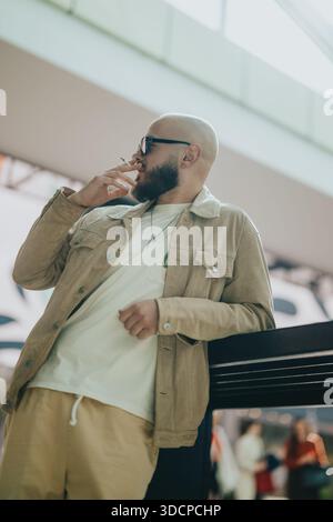 Homme occasionnel avec une barbe fumant une cigarette tout en se penchant sur la balustrade dans un cadre extérieur urbain Banque D'Images