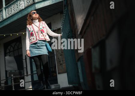 Femme en col roulé blanc, gilet folklorique, jupe en Jean et lunettes de soleil se tient à l'extérieur d'un bâtiment avec des balcons en bois bleu traditionnels. Banque D'Images