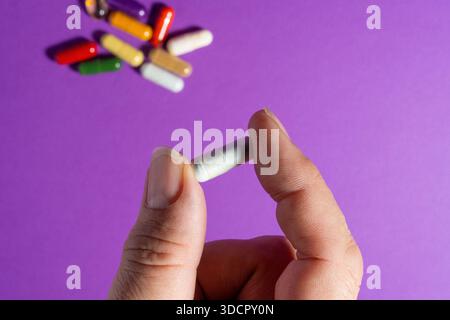 Gros plan d'une main tenant une pilule à capsule unique, plusieurs capsules colorées dispersées en arrière-plan sur la surface violette. Banque D'Images
