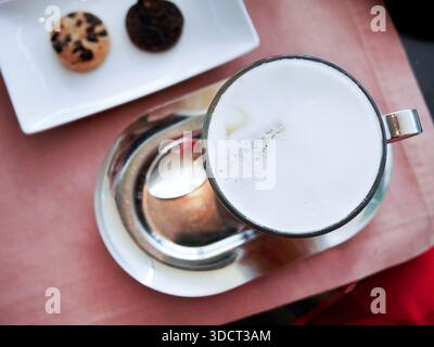 Photographie à plat représentant une tasse en verre de cappuccino chaud ou de latte recouverte d'une mousse de lait épaisse et crémeuse. Servi sur un plateau ovale argenté brillant avec une théière Banque D'Images
