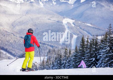 Vue arrière du skieur en équipement d'hiver lumineux avec bâtons et sac à dos sur la neige, surplombant la forêt alpine et les pistes de ski dans les montagnes Banque D'Images