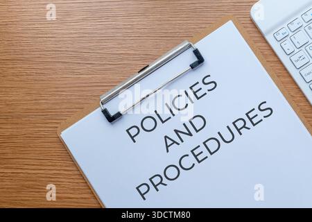 Un presse-papiers avec le texte « POLITIQUES ET PROCÉDURES » sur un bureau en bois à côté d'un clavier. Conformité de l'entreprise, réglementations commerciales, concept de directives. Banque D'Images