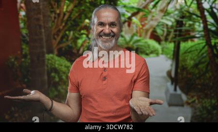 Homme aux longs cheveux gris et à la barbe tient les deux paumes sur un chemin de parc verdoyant dans la forêt, souriant doucement ; sérénité. Banque D'Images