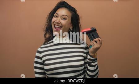 Femme tenant la manette de jeu vidéo et souriant dans le studio de pêche, haut rayé visible, montrant la main tatouée et boucle d'oreille de cerceau ; joie ludique. Banque D'Images