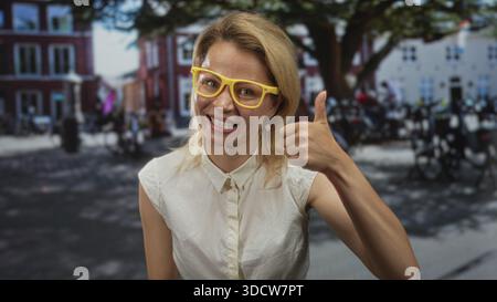 Jeune femme blonde souriante donne un geste du pouce vers le haut sur une rue urbaine animée en plein jour pendant la journée ensoleillée ; optimisme. Banque D'Images