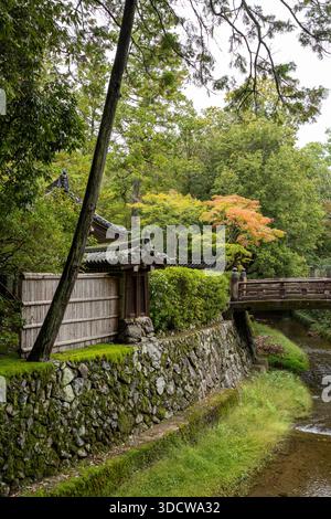 Un jardin japonais traditionnel avec une clôture en bambou, un mur de pierre et un pont en bois sur un ruisseau, entouré de verdure luxuriante et de feuillage d'automne. Banque D'Images