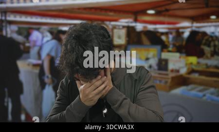 Jeune homme à la barbe riant à l'extérieur sur un marché de rue animé avec des gens flous et des stands colorés représentant une interaction sociale vibrante. Banque D'Images