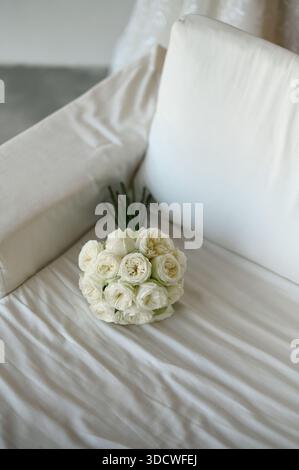 Un délicat bouquet de mariée blanc repose sur un canapé blanc doux, créant une scène sereine et minimaliste inspirée du mariage. Tons clairs, textures douces et ro Banque D'Images