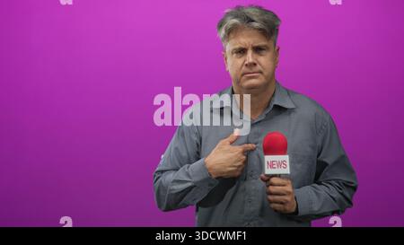 Journaliste homme d'âge moyen pointant le doigt sur la poitrine tout en tenant le microphone rouge étiqueté News contre le mur violet du studio ; hésitation. Banque D'Images
