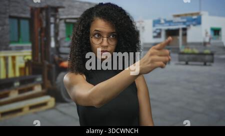Femme portant des lunettes se tient à l'extérieur dans une zone industrielle avec un chariot élévateur et des bâtiments en arrière-plan, regardant concentré et pointant. Banque D'Images
