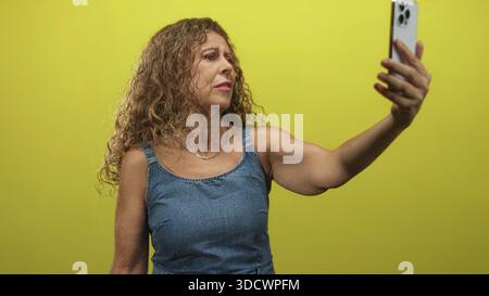 Femme étend le bras tenant smartphone pour selfie en studio, plissant et vérifiant son visage contre un mur jaune tout en portant un denim sans manches dres Banque D'Images