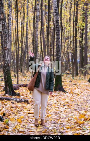 Femme souriante marche à travers une forêt d'automne, feuilles tombées jaunes sous les pieds, grands arbres, couleurs chaudes, humeur joyeuse et détendue Banque D'Images
