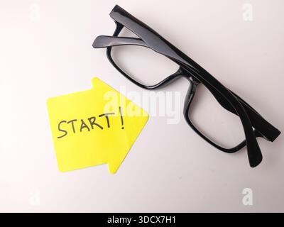 Note autocollante avec START ! écrit dessus à l'encre noire, pointant vers une paire de lunettes encadrées en noir. Banque D'Images
