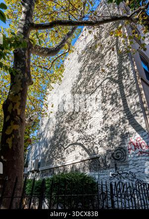 Motif d'ombre abstrait projeté sur le flanc d'un bâtiment à Chelsea, New York. Les branches et les feuilles des arbres créent des silhouettes superposées sur un mur texturé et altéré, mêlant formes naturelles et architecture urbaine. Les graffitis subtils et l'usure de surface ajoutent de la complexité visuelle, tandis que la lumière et le contraste forts soulignent la forme, le rythme et l'espace négatif. L'image explore l'intersection tranquille de la nature, du temps et de l'environnement bâti dans un cadre métropolitain dense. Banque D'Images