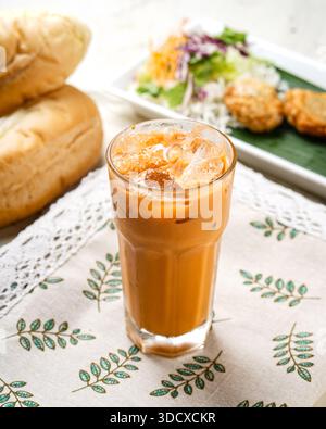 Un grand verre rempli de thé glacé thaïlandais crémeux et orange et de glaçons repose sur un tissu à motifs. En arrière-plan, des petits pains bruns dorés et une assiette Banque D'Images