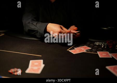 Mains de croupier de poker distribuant des cartes à jouer sur la table de casino sombre pendant le jeu de cartes avec des jetons, la surface feutrée, et l'éclairage de studio contrôlé, gros plan gamb Banque D'Images