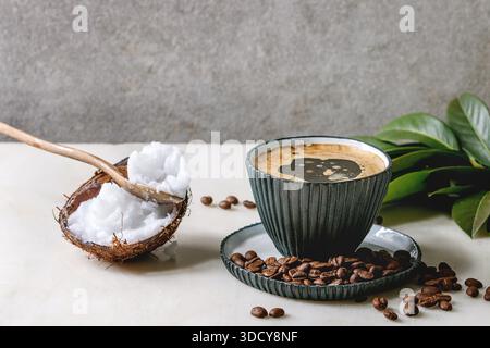 Café pare-balles. Café Keto Diet dans une tasse en céramique bleue avec de l'huile de presse à froid de noix de coco biologique dans des cuillères avec des haricots et branche verte sur une table en marbre blanc. Mur gris à l'arrière-plan. Banque D'Images
