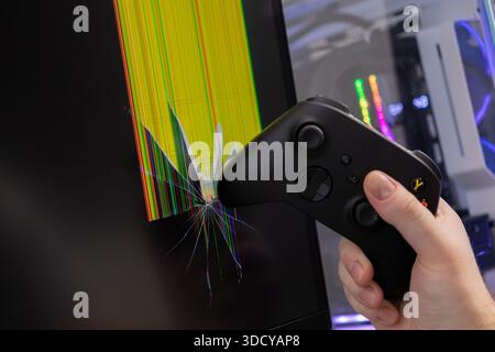 Gros plan de la main d'une personne tenant une manette de jeu noire frappant un moniteur d'ordinateur de bureau, provoquant un écran éclatant fissuré avec artefact numérique Banque D'Images