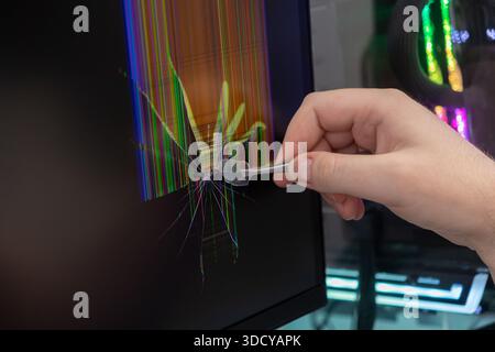 Une main utilise habilement une pince à épiler pour enlever un éclat de verre d'un écran de moniteur d'ordinateur gravement fissuré, affichant des lignes de dysfonctionnement colorées, indicati Banque D'Images