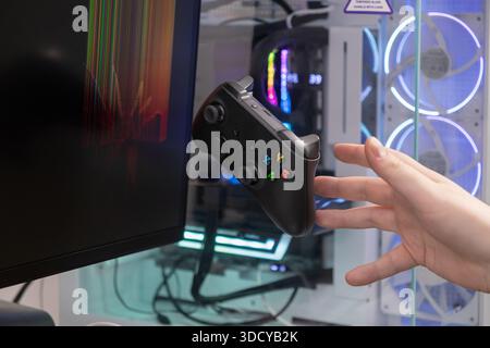 Gros plan d'une main tenant une manette de jeu sans fil noire à côté d'un écran d'ordinateur affichant des graphiques colorés et un puissant PC de jeu avec R. Banque D'Images